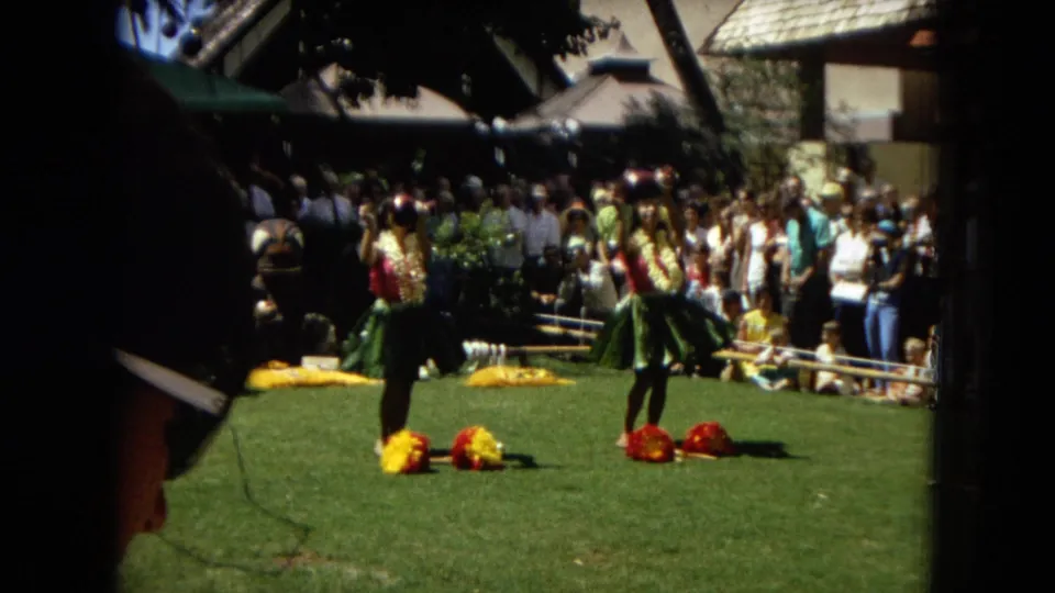 1967 Hawaii Super 8 frame showing hula dancers in motion on a lawn, with colorful flower props at their feet and spectators gathered closely behind.
