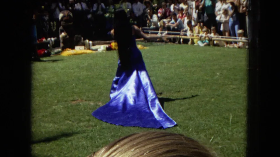 1967 Hawaii Super 8 frame showing a dancer in a blue skirt or dress crossing the grassy performance area, with spectators and lawn seating in the background.