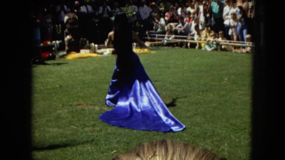 1967 Hawaii Super 8 frame showing a hula dancer in a long blue dress on grass, with spectators gathered behind the performance area.