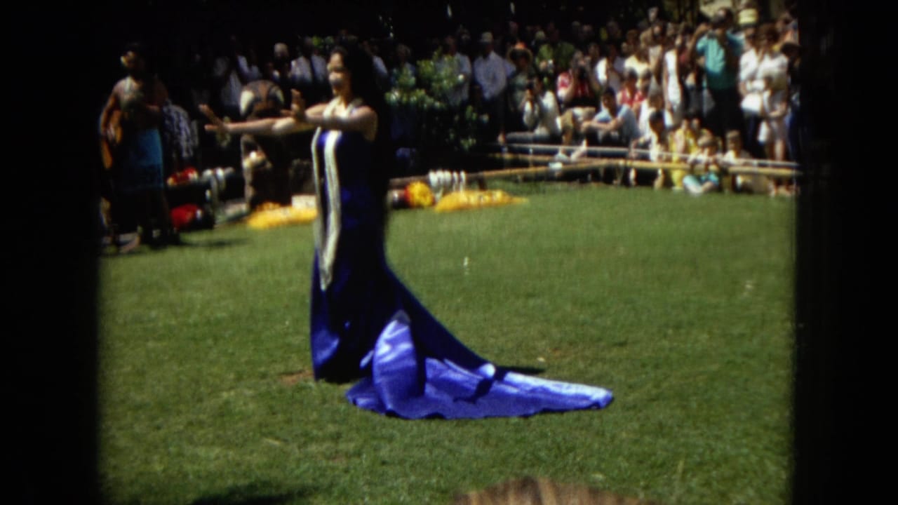 Hula dancer gestures with both arms while a crowd watches from the edge of a lawn.