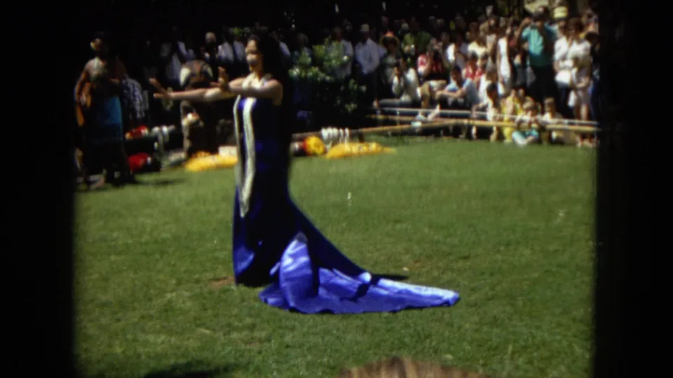 1967 Hawaii Super 8 frame with a hula performer in blue extending her arms on a grassy lawn as an audience watches behind her.