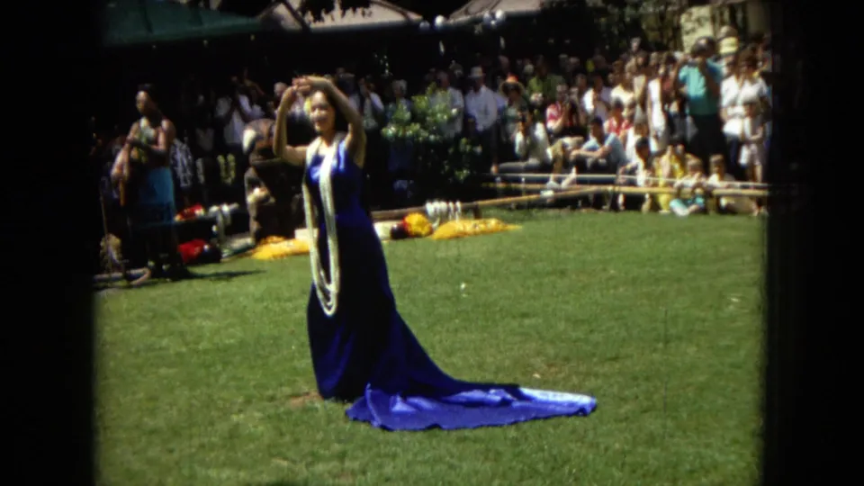 1967 Hawaii Super 8 frame showing a hula dancer raising her arms, with a musician and outdoor spectators in the background.