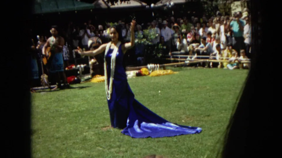 1967 Hawaii Super 8 frame with a hula dancer in a blue dress and lei, a guitarist at left, and a crowd watching from the background.