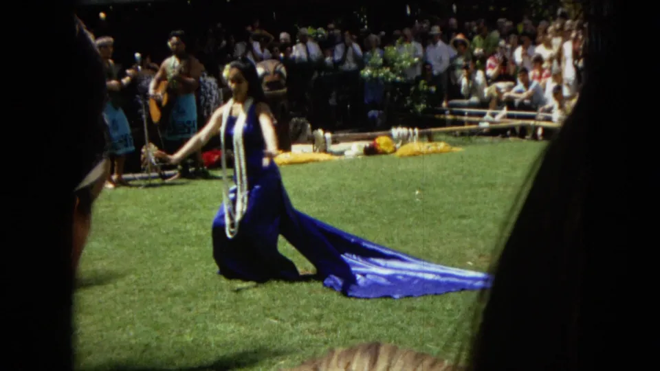 In 1967 Honolulu Super 8 footage, a woman wearing leis and a long blue dress kneels on a lawn during a hula performance with spectators and musicians behind her.