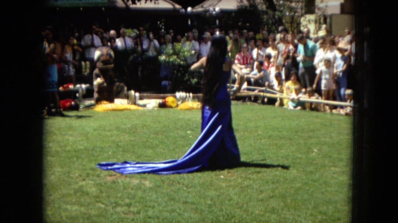 Woman in a long blue dress faces away while performing on grass before a crowd.