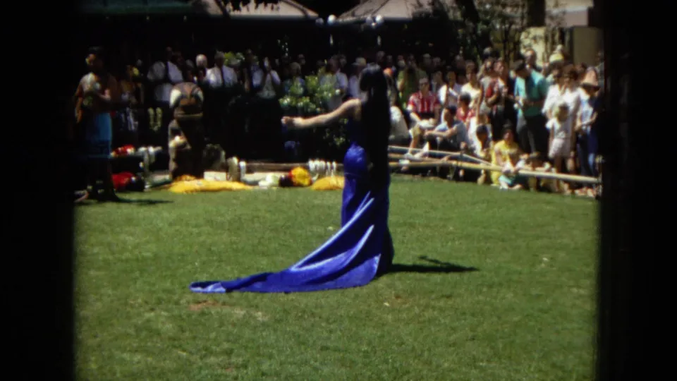 A woman in a blue dress extends her arm during a 1967 Honolulu hula performance, with musicians, props, and a crowd visible beyond the lawn.