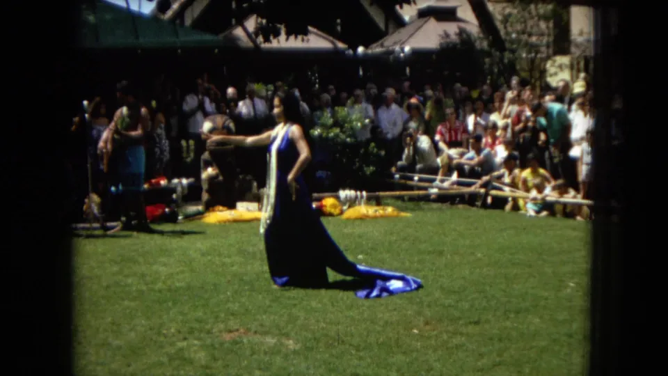 The performer stands in profile with one arm extended during the Honolulu hula scene, framed by a lawn, musicians, instruments, and a watching audience.