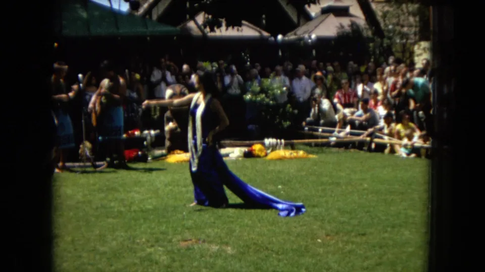 1967 Hawaii Super 8 frame showing a hula dancer in blue on grass, with musicians, microphones, and a crowded audience in the background.