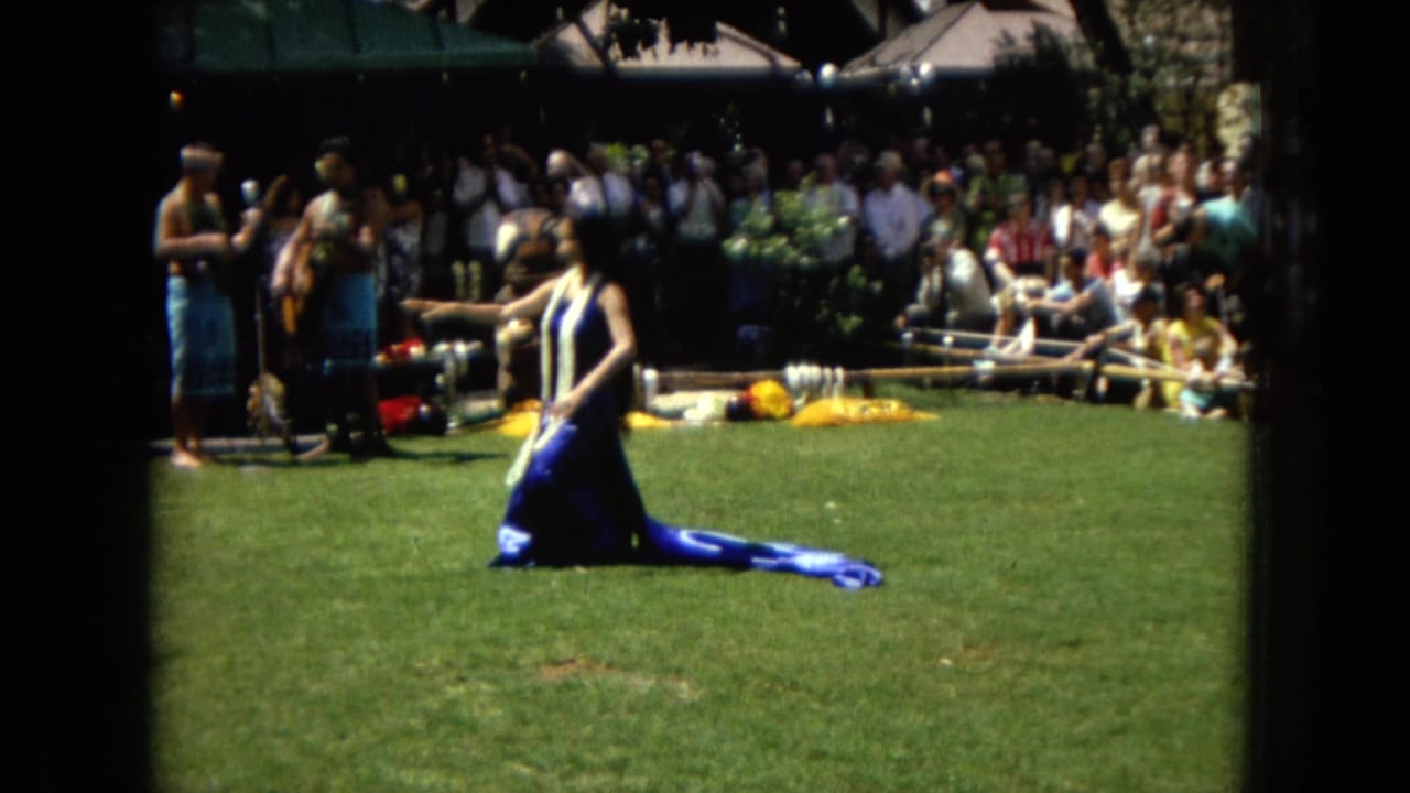1967 Hawaii Super 8 frame with a hula dancer moving across a sunlit lawn as musicians and spectators watch from the shaded background.