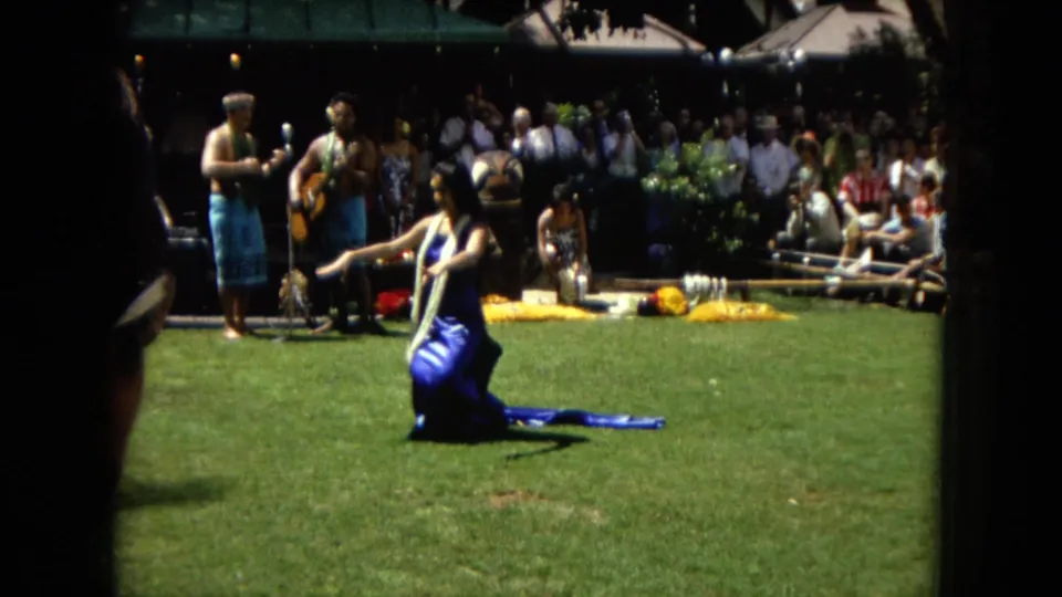 1967 Hawaii Super 8 frame showing a hula dancer kneeling and gesturing on grass, backed by live musicians and a closely gathered audience.