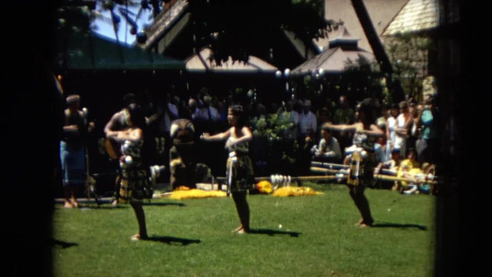 1967 Hawaii Super 8 frame showing two hula dancers on a lawn with a guitarist, other performers, and spectators gathered behind them.