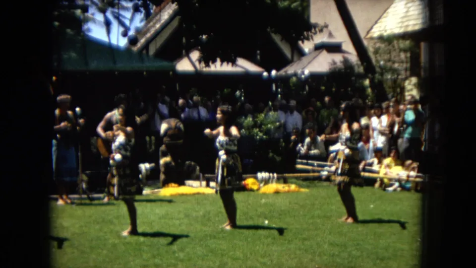 1967 Hawaii Super 8 frame showing three women performing hula on a sunny lawn while spectators and a band area sit in shade behind them.