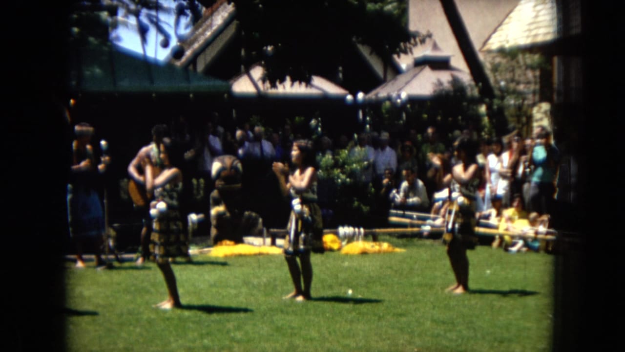1967 Hawaii Super 8 frame with hula dancers mid-performance, a shaded crowd behind them, and instruments or performers partly visible near the back of the lawn.