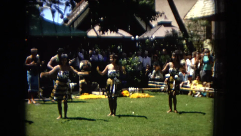 1967 Hawaii Super 8 frame showing three hula dancers facing the camera during an outdoor performance, with spectators, shade, and musical equipment behind them.