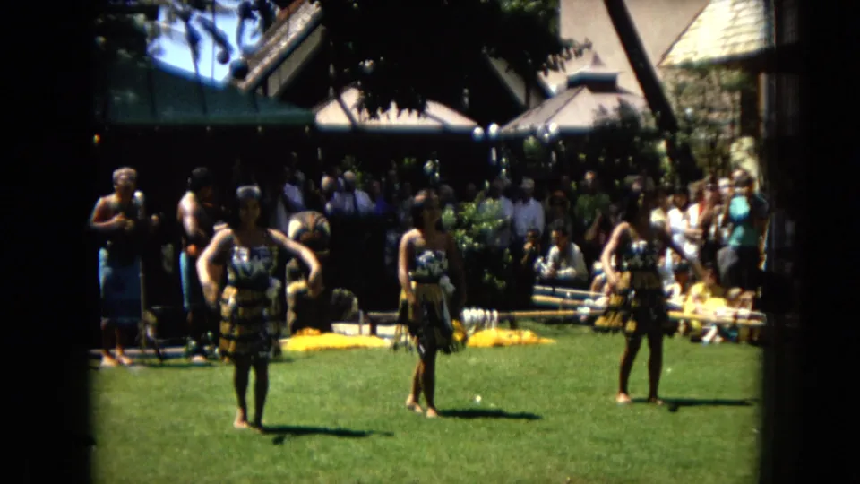 1967 Hawaii Super 8 frame showing hula dancers stepping across a sunlit lawn while a mixed audience watches from the shaded area behind them.