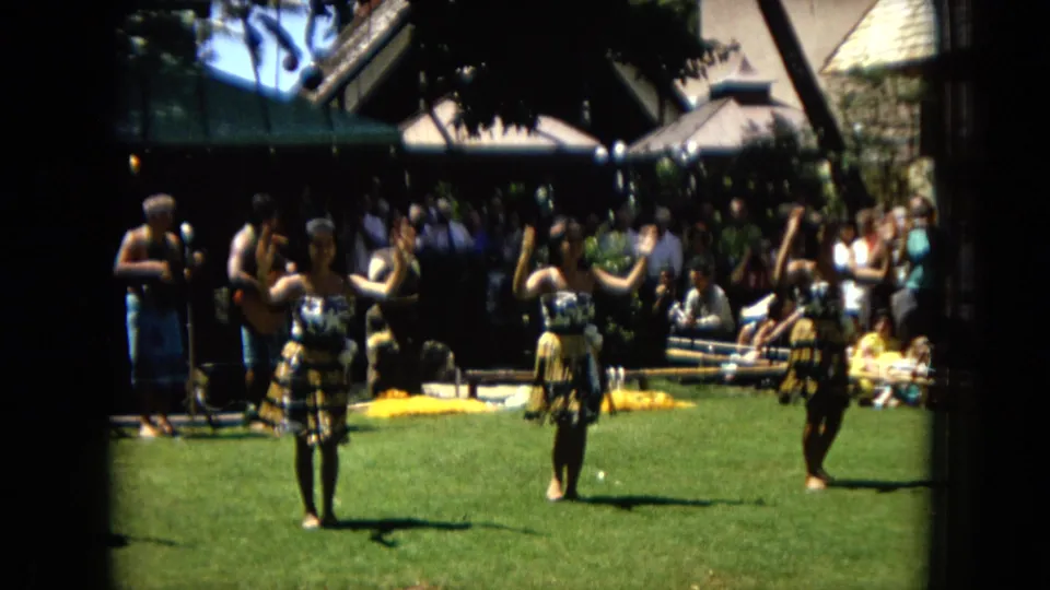 1967 Hawaii Super 8 frame showing women performing hula on a green lawn while musicians play and a crowd watches in the background.