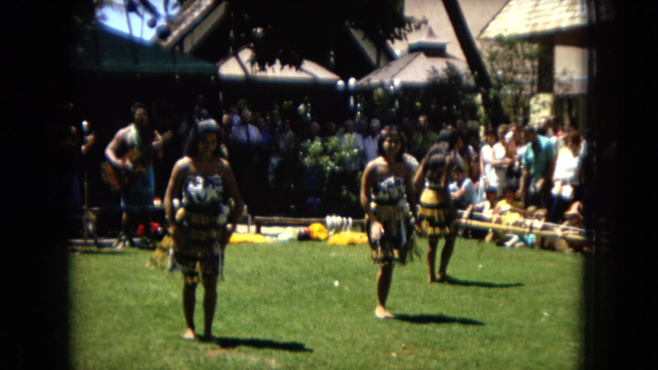 Three hula dancers stand on grass with a band and audience in the background.