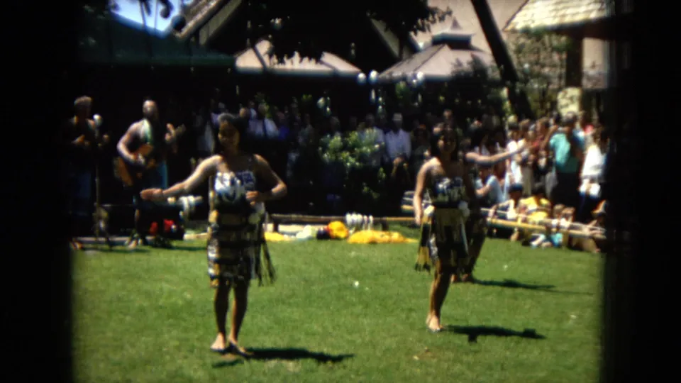 1967 Hawaii Super 8 still of two hula dancers closer to camera, performing arm movements on a lawn with musicians and spectators behind them.