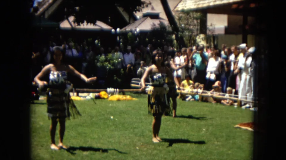 1967 Hawaii home-movie frame showing two hula dancers in mid-performance on a lawn, with a standing audience, musicians, and resort-like structures behind them.