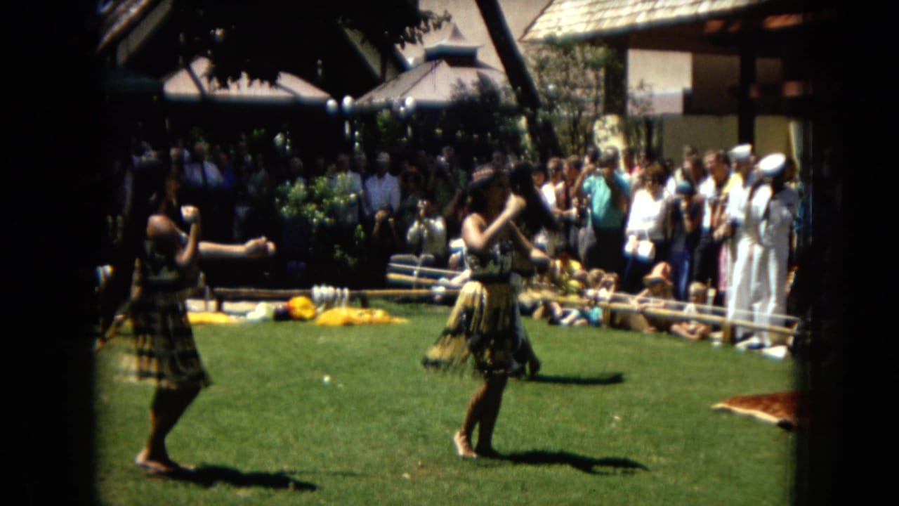 1967 Honolulu Super 8 frame showing women hula dancing outdoors on grass while tourists and seated spectators watch from behind a low barrier.