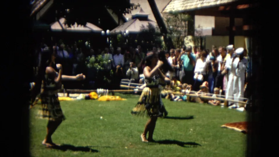 1967 Honolulu Super 8 frame showing women hula dancing outdoors on grass while tourists and seated spectators watch from behind a low barrier.