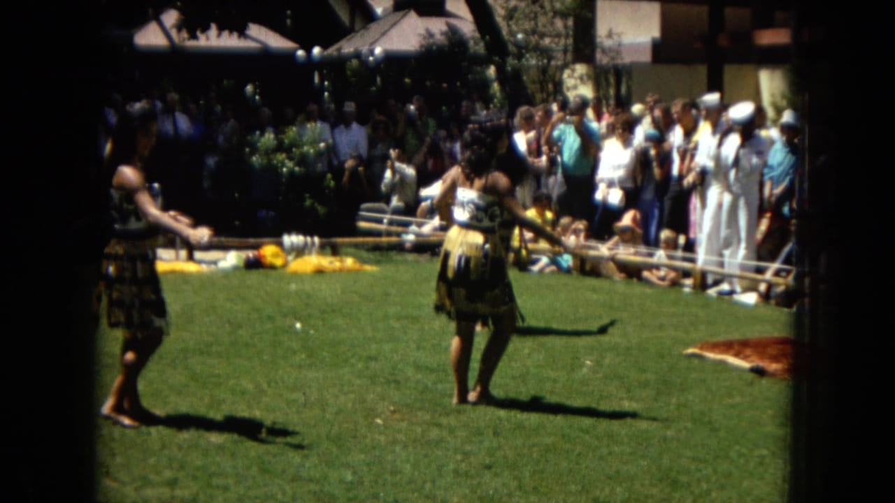 A hula dancer stands on the grass with musicians and spectators behind her.