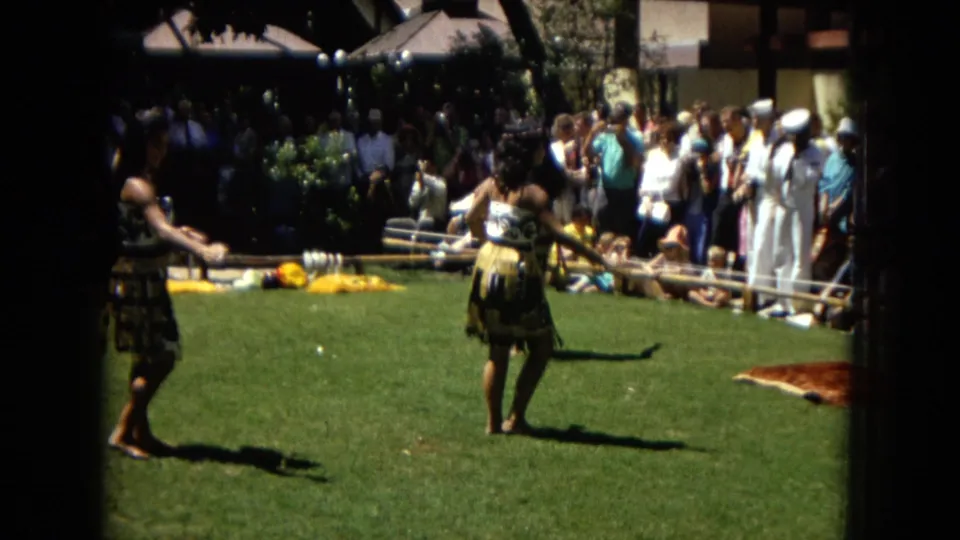 1967 Honolulu Super 8 frame with a hula dancer in a skirted costume on a lawn, backed by musicians, tourists, and low outdoor structures.