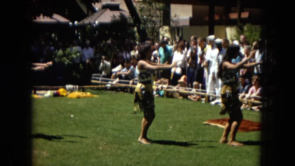 1967 Honolulu Super 8 frame showing two hula dancers in motion during an outdoor performance, with tourists and seated viewers gathered close to the lawn.