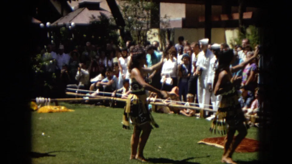 1967 Honolulu Super 8 frame showing a hula dancer near musicians at the edge of an outdoor lawn performance, with spectators gathered in the background.