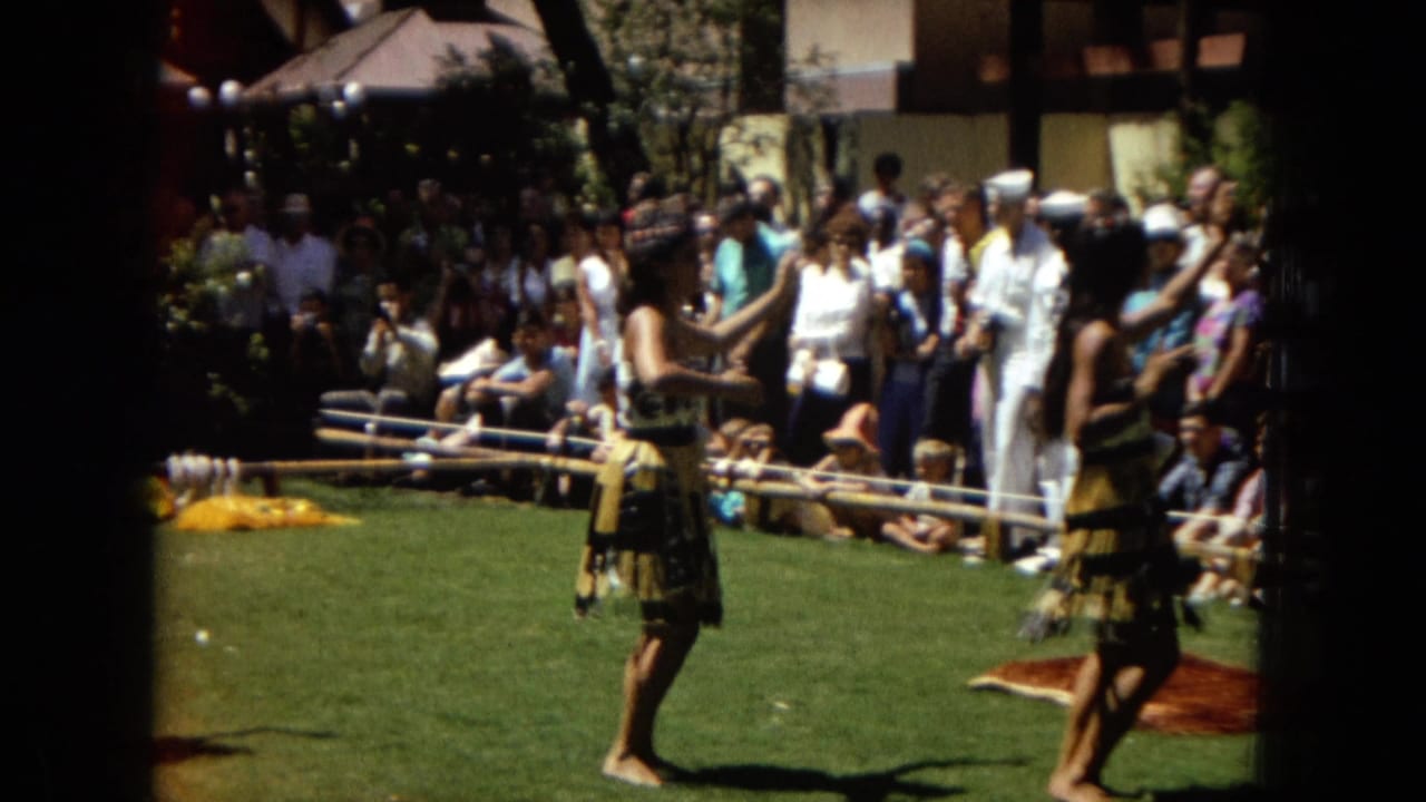 In 1967 Hawaii home-movie footage, two hula dancers perform on a grassy outdoor area while spectators watch from behind a low barrier.