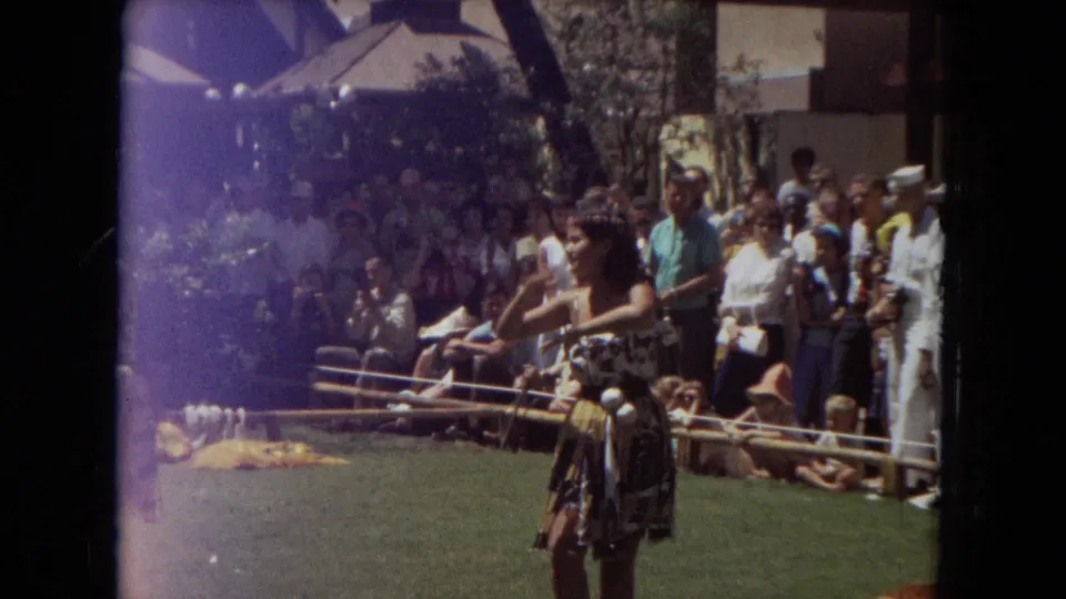 A hula dancer in patterned costume gestures during an outdoor Honolulu-area performance, with a crowd and shaded buildings visible behind her.