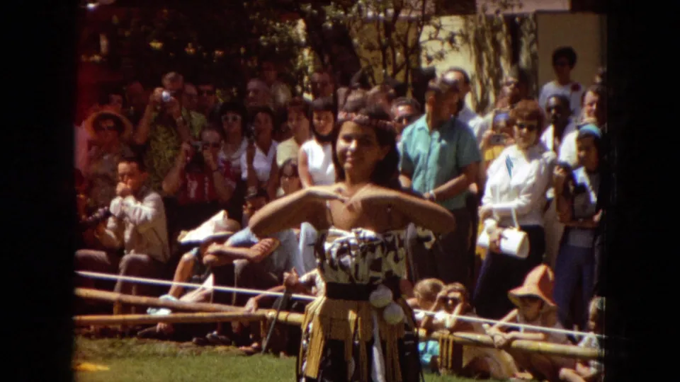 A closer Super 8 frame shows a smiling hula dancer in costume performing arm movements before a dense audience in 1967 Hawaii footage.