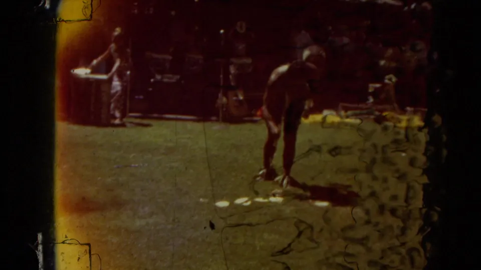 A darker frame with visible film edge wear shows a hula dancer on the grass while musicians or band members stand behind the performance area.