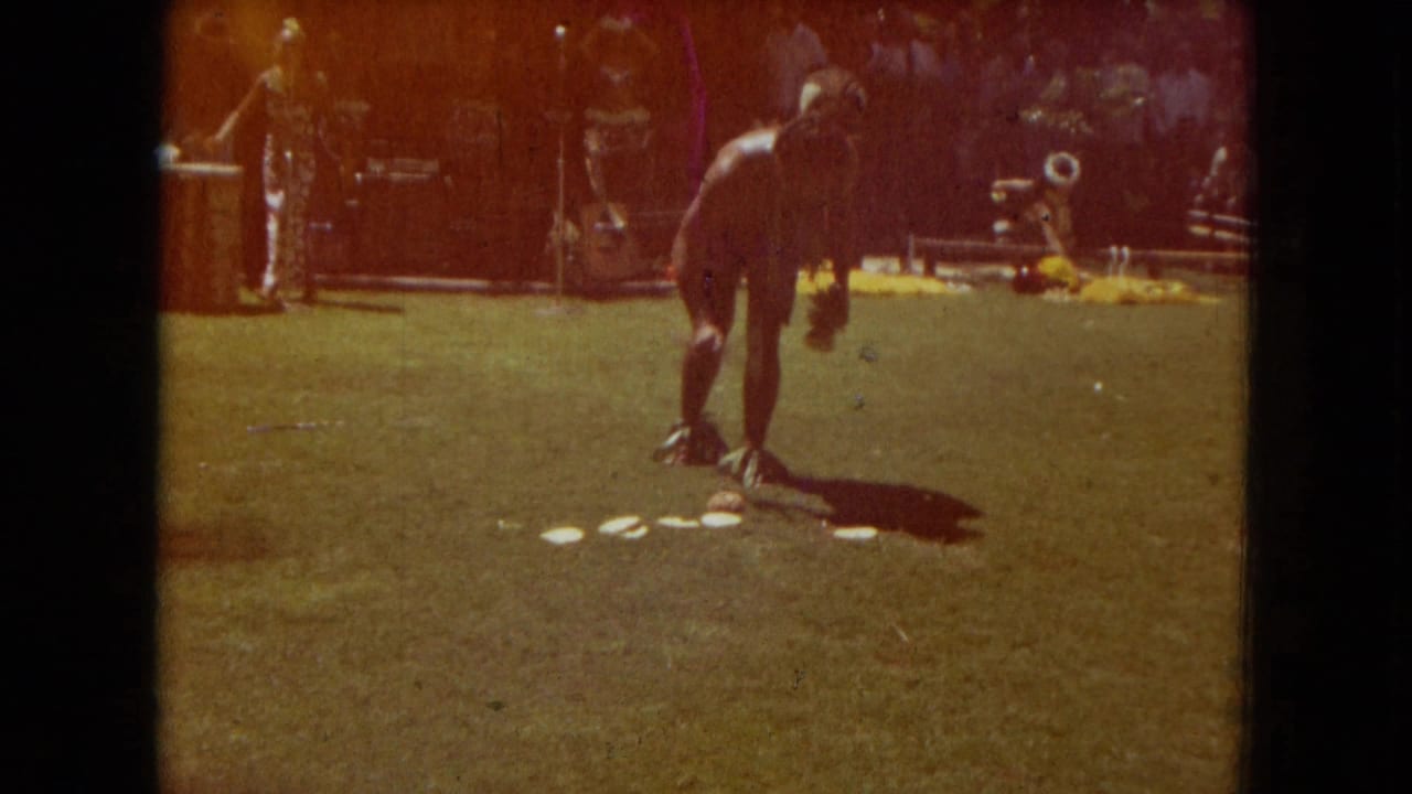 A 1967 Hawaii Super 8 frame shows a hula dancer bending or stepping on a sunny lawn while a small band and audience remain in the background.