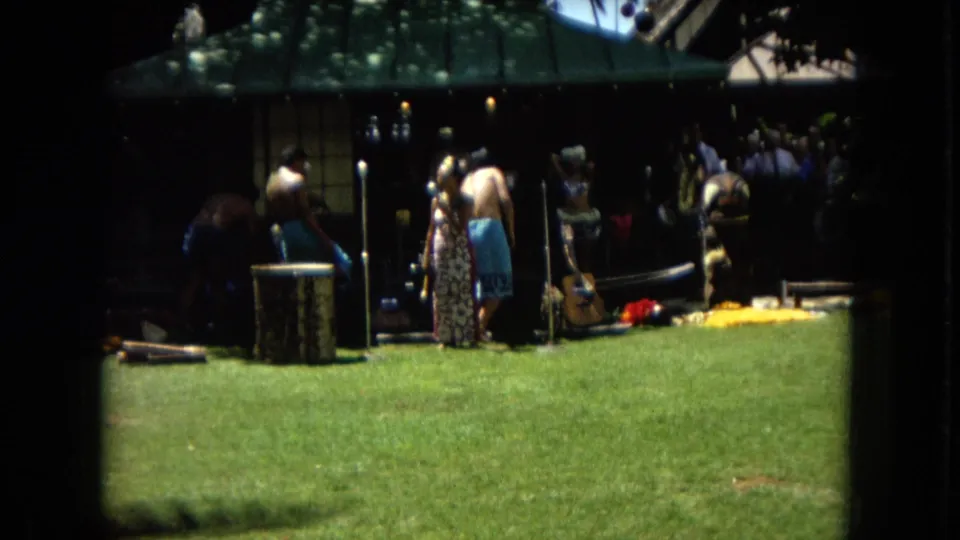 A Super 8 frame from the Hawaii performance shows musicians, performers, and spectators clustered near a shaded pavilion beside a bright green lawn.