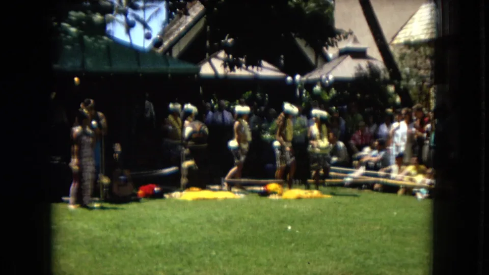 The frame shows a 1967 Hawaii outdoor hula performance area with a crowd watching from beside shaded structures and performers near the lawn.