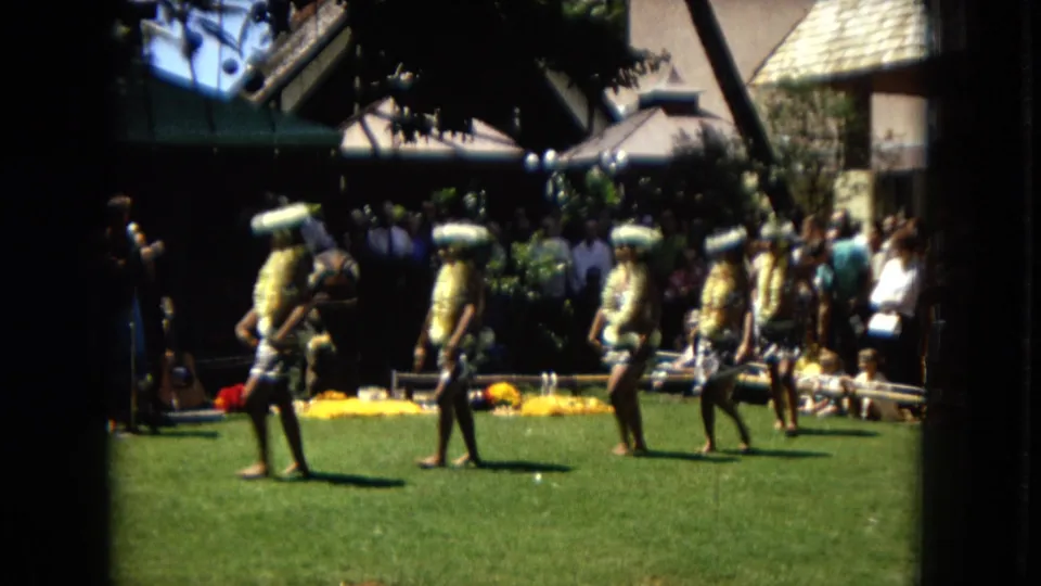 A 1967 Hawaii Super 8 frame shows several hula dancers in coordinated outfits moving across a sunlit lawn with spectators and musicians behind them.