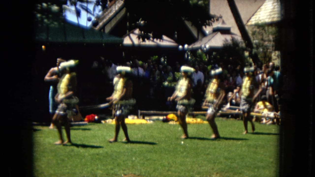 In 1967 Hawaii footage, women wearing leis and hula-style skirts perform outdoors on a lawn with musicians or attendants and seated spectators behind them.