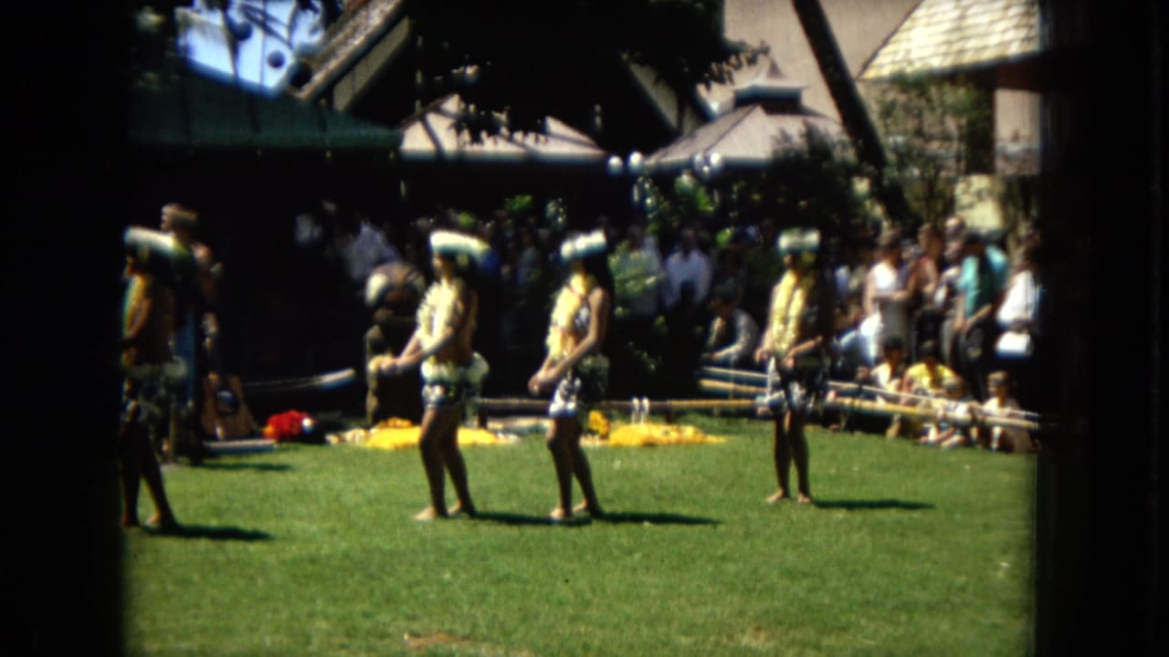 Dancers stand spaced across a sunny lawn with spectators and performers gathered behind them.