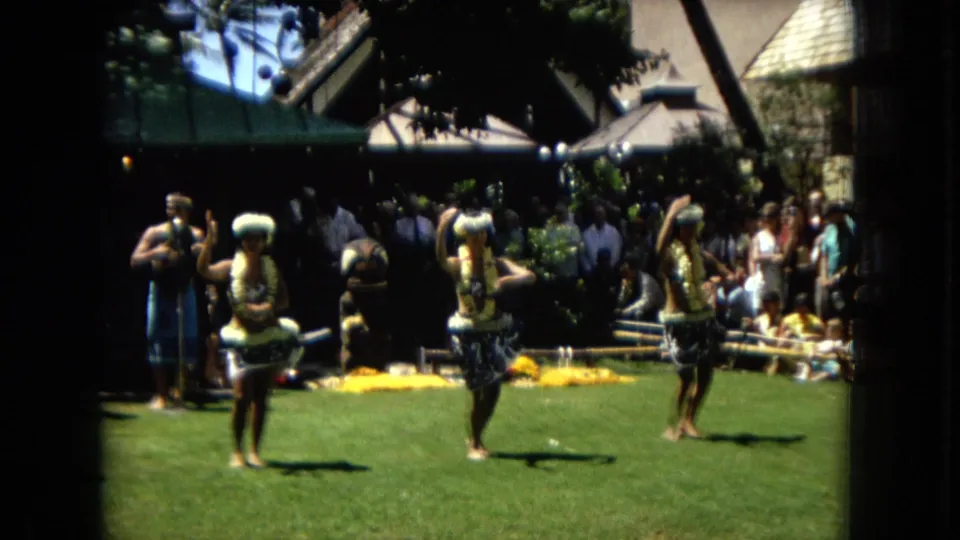 A closer Super 8 frame shows two hula dancers mid-routine on a lawn in 1967 Hawaii, with a standing man, spectators, and yellow flowers behind them.