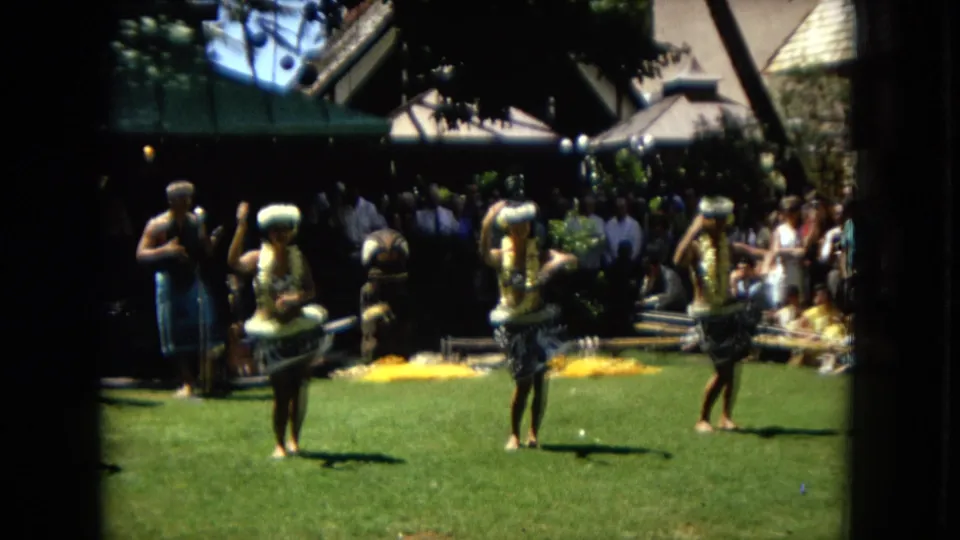 Three women in leis and hula-style skirts perform outdoors in 1967 Hawaii, framed by spectators, musicians or attendants, flower arrangements, and resort-like architecture.