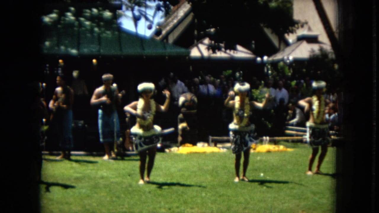 1967 Hawaii Super 8 frame showing women in leis and patterned skirts dancing hula outdoors with a small band and spectators in the background.