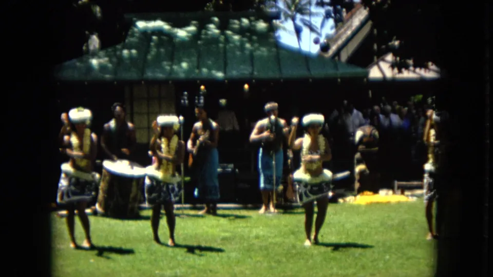 1967 Hawaii frame with hula dancers in floral adornments performing on a sunlit lawn as a drummer and musicians play behind them.