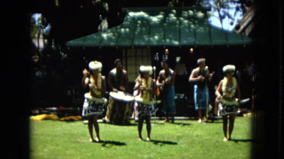 1967 Hawaii Super 8 still showing three women dancing hula in front of a shaded band area and seated onlookers.