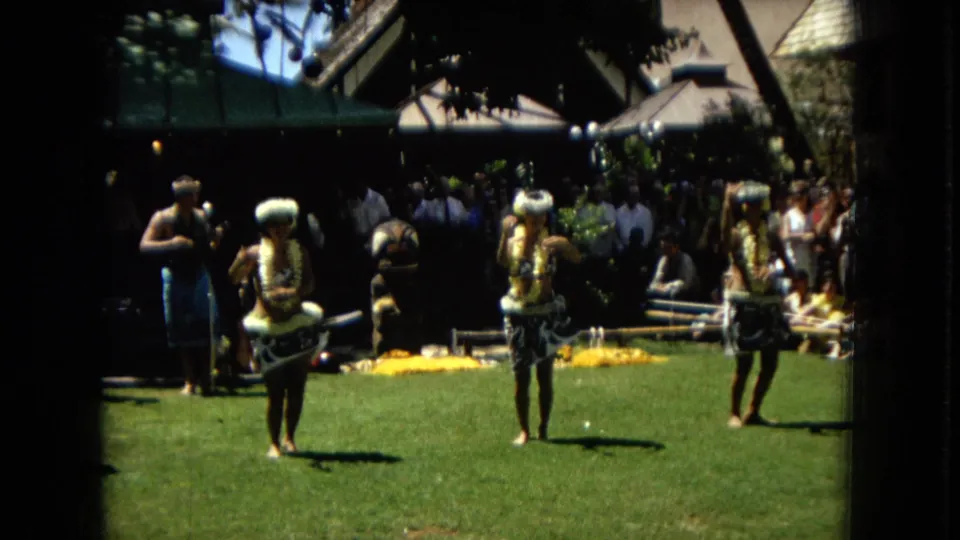1967 Hawaii frame of hula dancers in leis and patterned skirts performing outdoors, with musicians and spectators near a shaded structure.