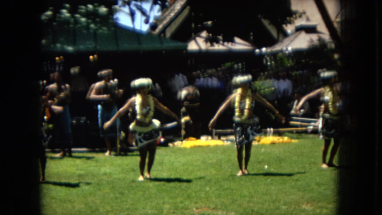 1967 Super 8 frame from Hawaii showing hula dancers in leis and headpieces performing on grass while musicians stand behind them.