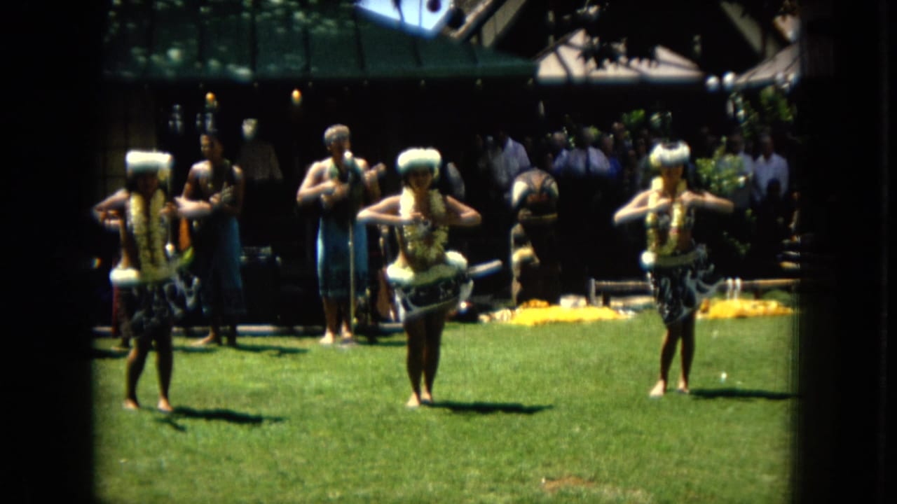 Three dancers move in formation on a grass lawn as a band plays behind them.