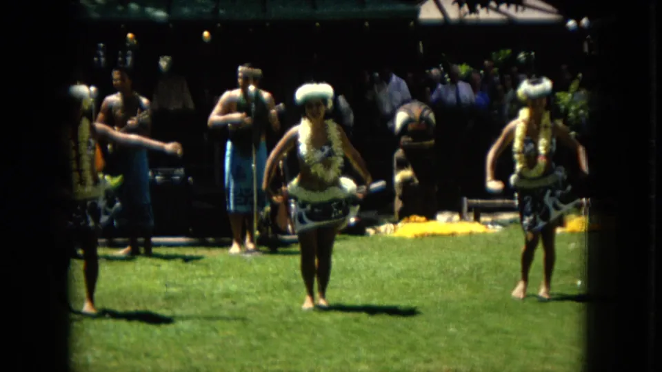 1967 Super 8 frame from Hawaii with hula dancers mid-routine, musicians behind them, and spectators partly visible in shade.