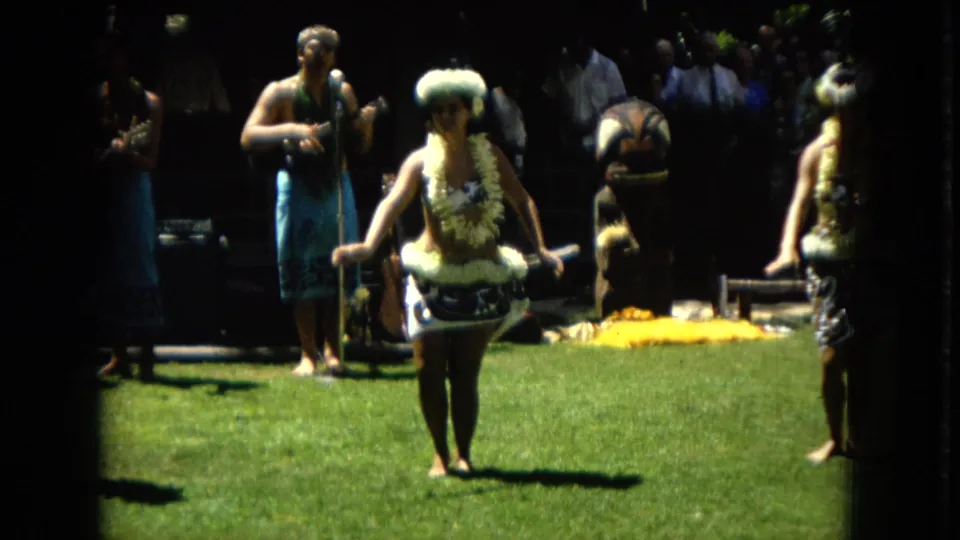 1967 Hawaii Super 8 frame showing a hula dancer centered on a sunlit lawn with a musician and other performers partly visible behind her.