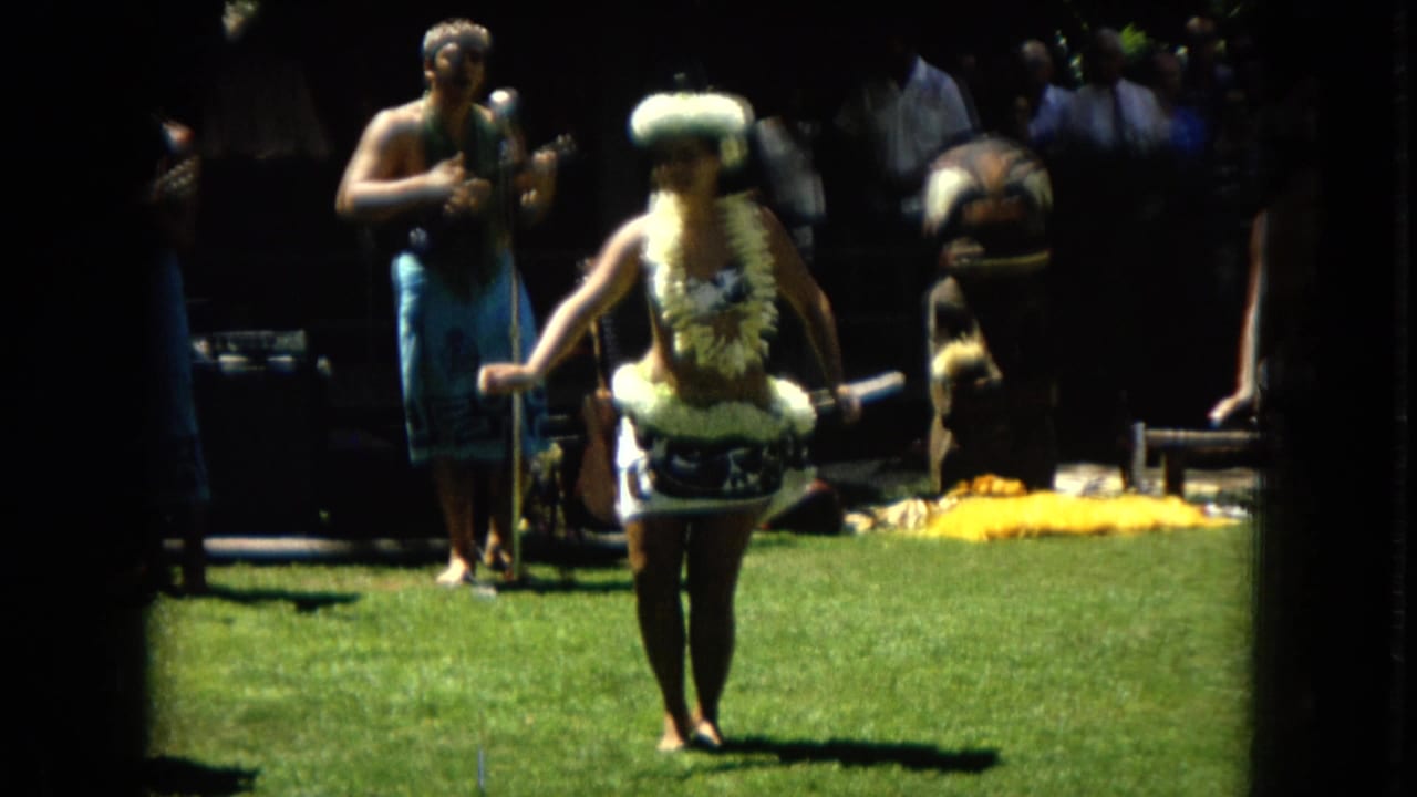 1967 Hawaii Super 8 frame showing a hula dancer wearing a floral lei and skirt accent on a grassy outdoor performance area with musicians behind her.